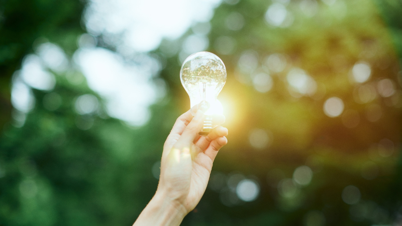 Holding a light bulb in one hand in the air, the bulb emits a warm light under the sunlight.